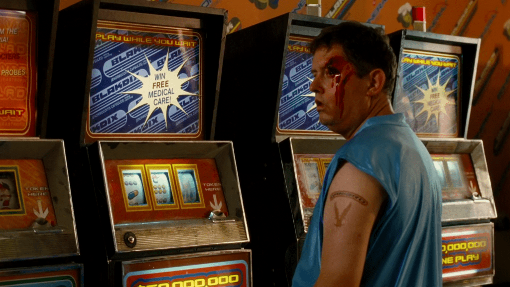 He sees a man with a blank expression and a bleeding head wound above his left eye, who stands a bank of slot machines. The backglass of each has diagonal logos advertising BLAKDIX capsules (n.b. the wallpaper advertises BONERAX), telling players they can play while they wait, and that they can WIN FREE MEDICAL CARE. The reel strips don’t show bells or fruit, but rather, pills. The blood from the head wound shines in the lights.